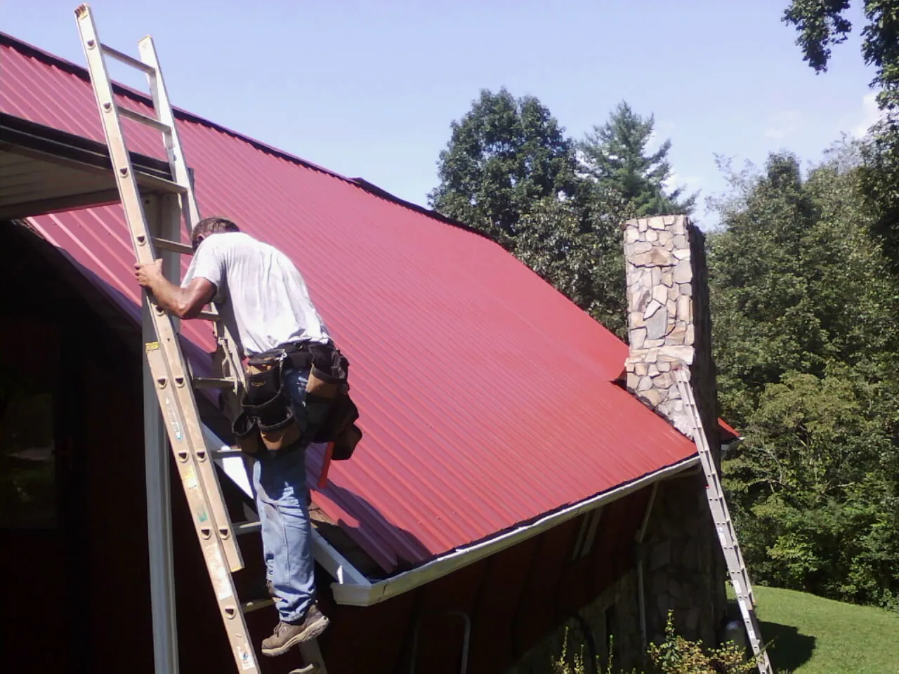 Roofer on a ladder beside a red metal roof and stone chimney — Soffit & Fascia Repair by Roofing Brothers in Cascade